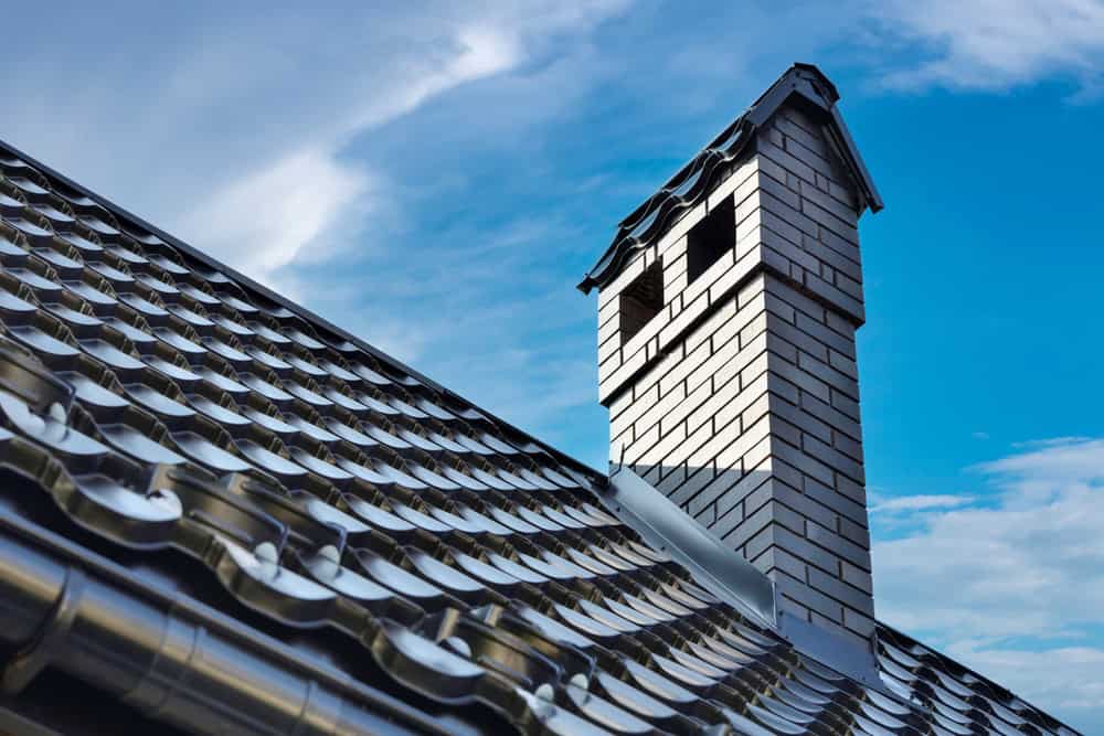 Close-up of a house rooftop with a shiny metallic tile roof and a white brick chimney under a blue sky with scattered clouds. The chimney, which appears to need chimney repair, has two small rectangular openings near the top.