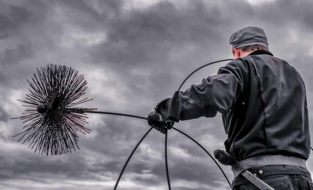 A chimney sweep dressed in dark, heavy clothing and a cap holds a large round brush attached to a pole against a backdrop of a cloudy, overcast sky. He appears to be preparing for fireplace cleaning.