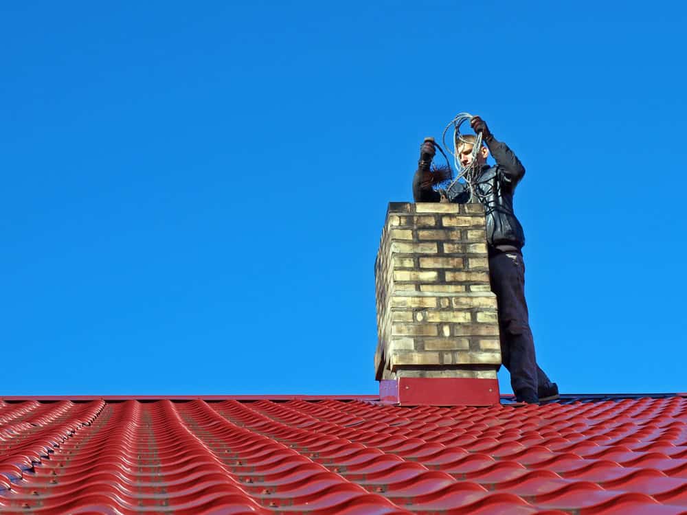 A person dressed in black winter clothing is standing next to a chimney on a red corrugated metal roof under a clear blue sky. They appear to be performing chimney services, handling a coiled wire or cable while adjusting something on the chimney with their other hand.